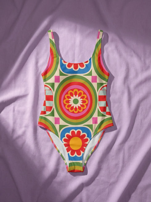 A one-piece swimsuit with a 1970s retro psychedelic geometric pattern in red, orange, green, and blue on a cream background. The swimsuit features a scoop neck and cheeky high-cut fit. In one view, she styles it with a pink baseball cap and gold necklaces. The design includes a central radial sunburst mandala and checkerboard side panels, representing a maximalist, rockstar-ready aesthetic.