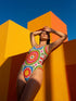 A one-piece swimsuit with a 1970s retro psychedelic geometric pattern in red, orange, green, and blue on a cream background. The swimsuit features a scoop neck and cheeky high-cut fit. In one view, she styles it with a pink baseball cap and gold necklaces. The design includes a central radial sunburst mandala and checkerboard side panels, representing a maximalist, rockstar-ready aesthetic.