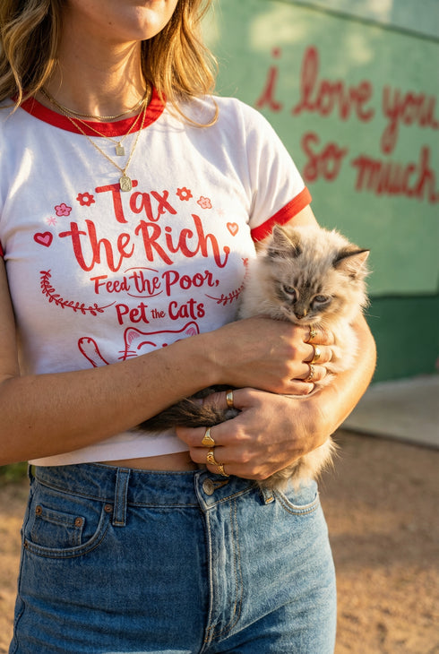 White women's cropped ringer t-shirt with bright red trim on the collar and sleeves. The chest features a cute kawaii graphic in red and soft pink. The text reads "Tax the Rich" in a retro serif font, followed by "Feed the Poor" and "Pet the Cats" in script and sans-serif. Surrounding the text are hearts, sparkles, and flowers. At the bottom, a happy line-art cat with closed smiling eyes raises its paws in the air. The style is flat, 2D, and anime-inspired.