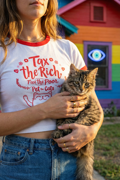 White women's cropped ringer t-shirt with bright red trim on the collar and sleeves. The chest features a cute kawaii graphic in red and soft pink. The text reads "Tax the Rich" in a retro serif font, followed by "Feed the Poor" and "Pet the Cats" in script and sans-serif. Surrounding the text are hearts, sparkles, and flowers. At the bottom, a happy line-art cat with closed smiling eyes raises its paws in the air. The style is flat, 2D, and anime-inspired.