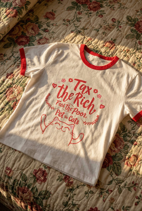 White women's cropped ringer t-shirt with bright red trim on the collar and sleeves. The chest features a cute kawaii graphic in red and soft pink. The text reads "Tax the Rich" in a retro serif font, followed by "Feed the Poor" and "Pet the Cats" in script and sans-serif. Surrounding the text are hearts, sparkles, and flowers. At the bottom, a happy line-art cat with closed smiling eyes raises its paws in the air. The style is flat, 2D, and anime-inspired.