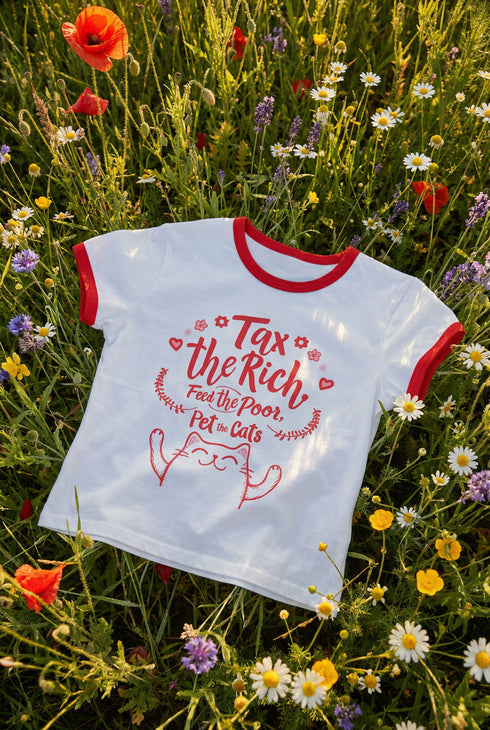 White women's cropped ringer t-shirt with bright red trim on the collar and sleeves. The chest features a cute kawaii graphic in red and soft pink. The text reads "Tax the Rich" in a retro serif font, followed by "Feed the Poor" and "Pet the Cats" in script and sans-serif. Surrounding the text are hearts, sparkles, and flowers. At the bottom, a happy line-art cat with closed smiling eyes raises its paws in the air. The style is flat, 2D, and anime-inspired.