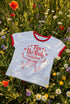 White women's cropped ringer t-shirt with bright red trim on the collar and sleeves. The chest features a cute kawaii graphic in red and soft pink. The text reads "Tax the Rich" in a retro serif font, followed by "Feed the Poor" and "Pet the Cats" in script and sans-serif. Surrounding the text are hearts, sparkles, and flowers. At the bottom, a happy line-art cat with closed smiling eyes raises its paws in the air. The style is flat, 2D, and anime-inspired.