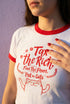 White women's cropped ringer t-shirt with bright red trim on the collar and sleeves. The chest features a cute kawaii graphic in red and soft pink. The text reads "Tax the Rich" in a retro serif font, followed by "Feed the Poor" and "Pet the Cats" in script and sans-serif. Surrounding the text are hearts, sparkles, and flowers. At the bottom, a happy line-art cat with closed smiling eyes raises its paws in the air. The style is flat, 2D, and anime-inspired.