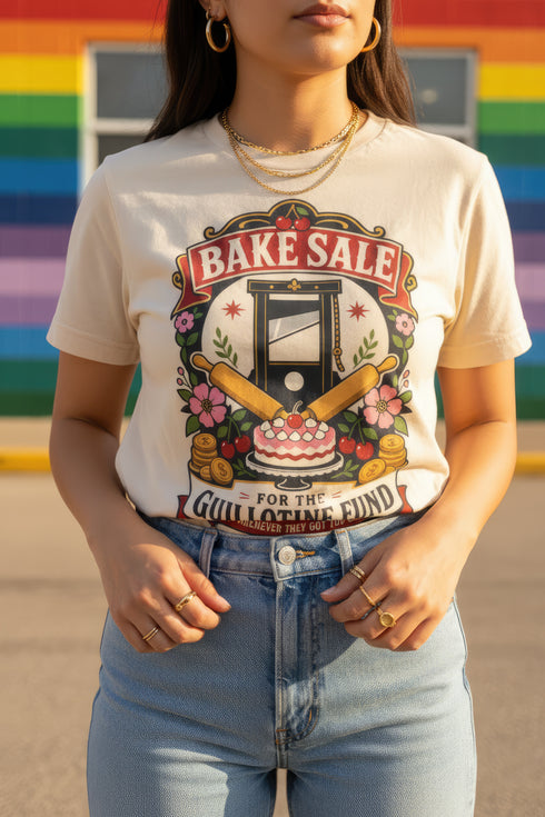 A short sleeve unisex crewneck t-shirt featuring a vintage style emblem graphic centered on the chest. The design features a red banner reading BAKE SALE above a black guillotine. Two crossed wooden rolling pins sit below the guillotine, above a layered pink and white cake with a cherry. The graphic is flanked by pink flowers, cherries, and stacks of gold coins. A bottom banner reads FOR THE GUILLOTINE FUND EST. WHENEVER THEY GOT TOO GREEDY. The shirt comes in light pink, cream, and blue.