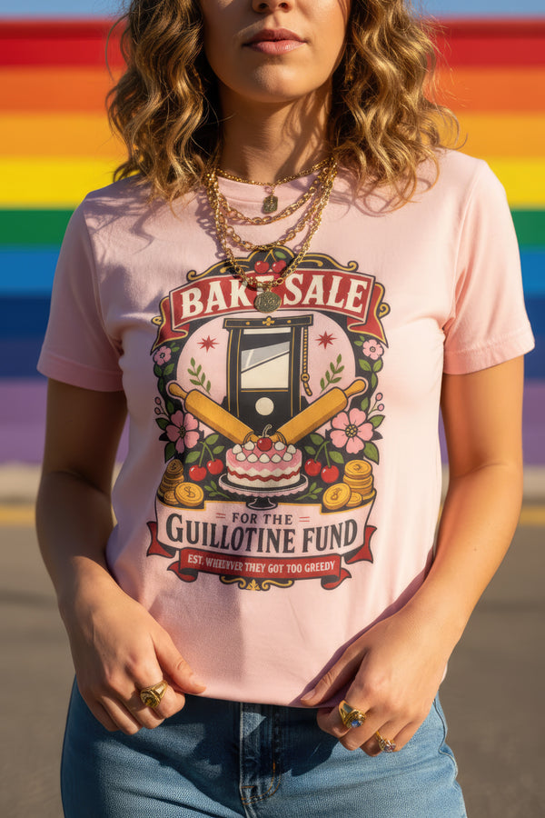 A short sleeve unisex crewneck t-shirt featuring a vintage style emblem graphic centered on the chest. The design features a red banner reading BAKE SALE above a black guillotine. Two crossed wooden rolling pins sit below the guillotine, above a layered pink and white cake with a cherry. The graphic is flanked by pink flowers, cherries, and stacks of gold coins. A bottom banner reads FOR THE GUILLOTINE FUND EST. WHENEVER THEY GOT TOO GREEDY. The shirt comes in light pink, cream, and blue.