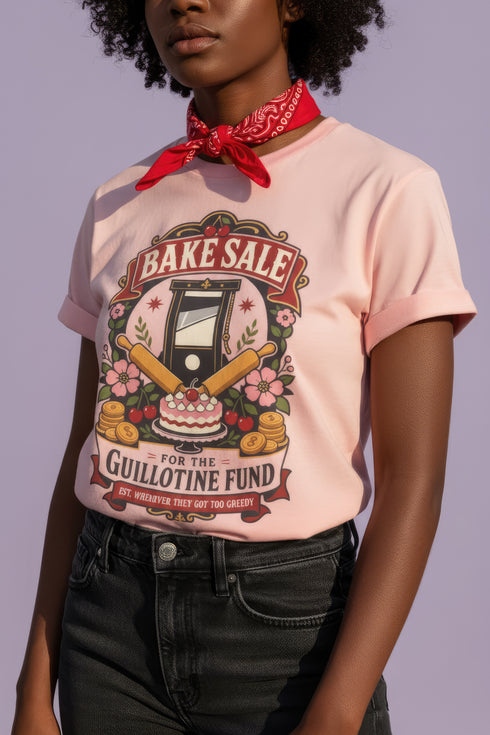 A short sleeve unisex crewneck t-shirt featuring a vintage style emblem graphic centered on the chest. The design features a red banner reading BAKE SALE above a black guillotine. Two crossed wooden rolling pins sit below the guillotine, above a layered pink and white cake with a cherry. The graphic is flanked by pink flowers, cherries, and stacks of gold coins. A bottom banner reads FOR THE GUILLOTINE FUND EST. WHENEVER THEY GOT TOO GREEDY. The shirt comes in light pink, cream, and blue.