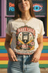 A short sleeve unisex crewneck t-shirt featuring a vintage style emblem graphic centered on the chest. The design features a red banner reading BAKE SALE above a black guillotine. Two crossed wooden rolling pins sit below the guillotine, above a layered pink and white cake with a cherry. The graphic is flanked by pink flowers, cherries, and stacks of gold coins. A bottom banner reads FOR THE GUILLOTINE FUND EST. WHENEVER THEY GOT TOO GREEDY. The shirt comes in light pink, cream, and blue.