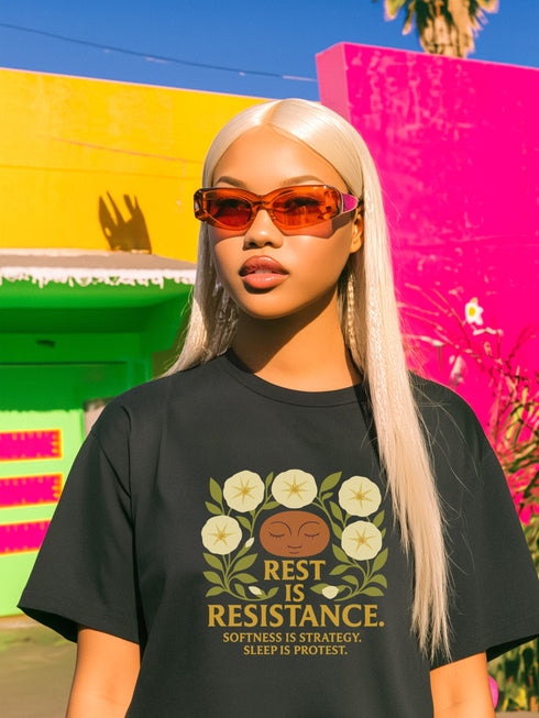 A black unisex t-shirt with a central front graphic. The design features the words "REST IS RESISTANCE" in a bold, gold, retro font. Above the main text, a serene, stylized face with closed eyes is framed by six cream-colored morning glory flowers and green leaves in a folk art, botanical style. Below, smaller text reads "SOFTNESS IS STRATEGY. SLEEP IS PROTEST." This alternative fashion, activist graphic tee is