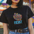 Black t-shirt with vibrant multicolored patterned raised fist graphic and blue "RESIST" text underneath. Rainbow activist protest shirt with psychedelic design elements on cotton-polyester blend.