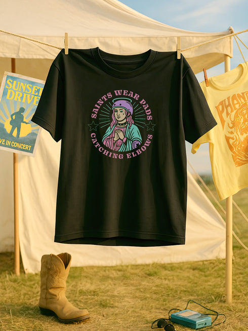 Black unisex t-shirt with bold graphic that reads "Saints Wear Pads Catching Elbows" surrounding a pastel saint figure wearing a roller skate helmet and protective pads. The design mimics embroidery in a retro tattoo-inspired art style. Available in sizes S to 3XL, this skate tee is soft, comfortable, and perfect for roller derby lovers, punk skaters, and feminist fashion fans. Great birthday or skate party gift idea.