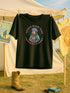 Black unisex t-shirt with bold graphic that reads "Saints Wear Pads Catching Elbows" surrounding a pastel saint figure wearing a roller skate helmet and protective pads. The design mimics embroidery in a retro tattoo-inspired art style. Available in sizes S to 3XL, this skate tee is soft, comfortable, and perfect for roller derby lovers, punk skaters, and feminist fashion fans. Great birthday or skate party gift idea.