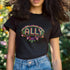 Black t-shirt featuring "ALLY" in gold letters protected by a rainbow arch and flanked by two sunflowers. LGBTQ+ pride ally shirt with colorful folk art-inspired design, available in cotton-poly blend, sizes S-3XL.