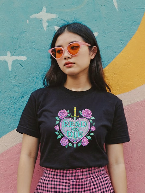 Unisex black t-shirt featuring a pastel goth “Read or Die” design in faux embroidery style. Centered heart motif pierced by a gold dagger, wrapped in pink roses and gothic script. Soft vintage-style tee made of cotton or cotton blend, perfect for book lovers, goth girls, alt fashion fans, and librarians with attitude. Great for dark academia, pastel punk, or cottagecore aesthetics. Ideal gift for readers, introverts, and anti-mainstream bibliophiles.