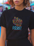 Black t-shirt with vibrant multicolored patterned raised fist graphic and blue "RESIST" text underneath. Rainbow activist protest shirt with psychedelic design elements on cotton-polyester blend.
