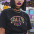 Black t-shirt featuring "ALLY" in gold letters protected by a rainbow arch and flanked by two sunflowers. LGBTQ+ pride ally shirt with colorful folk art-inspired design, available in cotton-poly blend, sizes S-3XL.