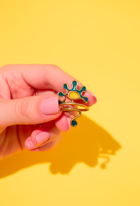 A close-up of a Berber Gripoix Wrap Ring in brass and enamel, featuring abstract teal and amber shapes on a yellow background. The ring’s sculptural wrap design and artistic details are highlighted in bright, natural light. Handmade statement jewelry by Honey Dazed.