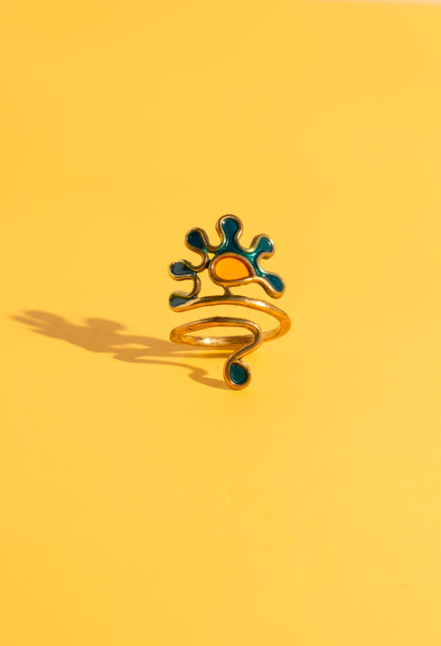 A close-up of a Berber Gripoix Wrap Ring in brass and enamel, featuring abstract teal and amber shapes on a yellow background. The ring’s sculptural wrap design and artistic details are highlighted in bright, natural light. Handmade statement jewelry by Honey Dazed.