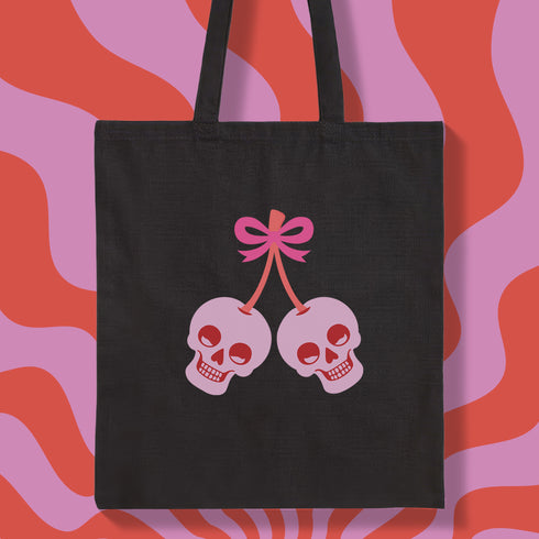 Canvas tote bag featuring pink skull cherries design. Two pink skulls connected by red stem and bow. Quirky gothic-inspired graphic on cream background. Stylish, edgy accessory for everyday use. Perfect for alternative fashion lovers.