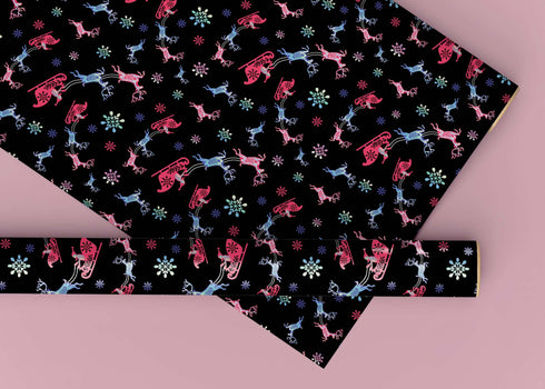 Christmas wrapping paper with a black background featuring colorful, stylized reindeer and Santa's sleigh designs. The reindeer and sleigh silhouettes are filled with skeletal patterns in shades of blue, pink, and red. Multicolored snowflakes in various sizes are scattered throughout. The paper has a whimsical, slightly gothic aesthetic that blends traditional holiday imagery with a unique, edgy twist.