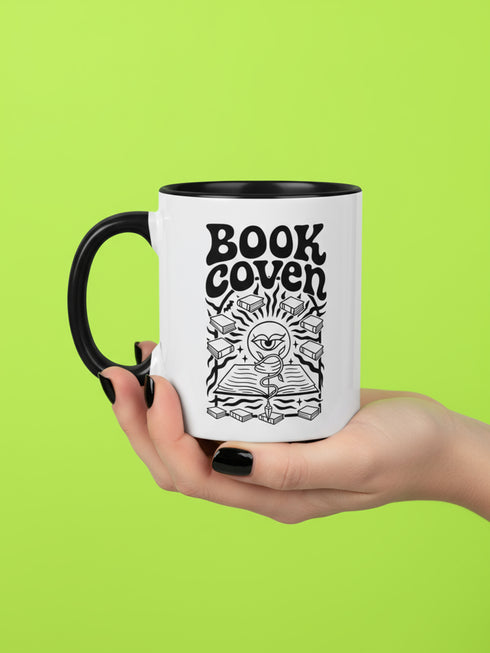 A two-tone ceramic mug with a glossy white exterior and a contrasting black interior, handle, and rim. The front of the mug features a black, rectangular graphic with the words "BOOK COVEN" in a bold, wavy, 70s-inspired retro font. Below the text is an occult-style illustration of an open book on a stand. An all-seeing eye with a snake coiled around it emerges from the book's pages, radiating lines of light. Smaller books float around the central image, completing the witchy, bookish design