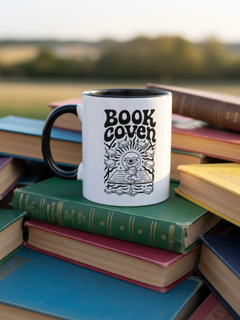 A two-tone ceramic mug with a glossy white exterior and a contrasting black interior, handle, and rim. The front of the mug features a black, rectangular graphic with the words "BOOK COVEN" in a bold, wavy, 70s-inspired retro font. Below the text is an occult-style illustration of an open book on a stand. An all-seeing eye with a snake coiled around it emerges from the book's pages, radiating lines of light. Smaller books float around the central image, completing the witchy, bookish design