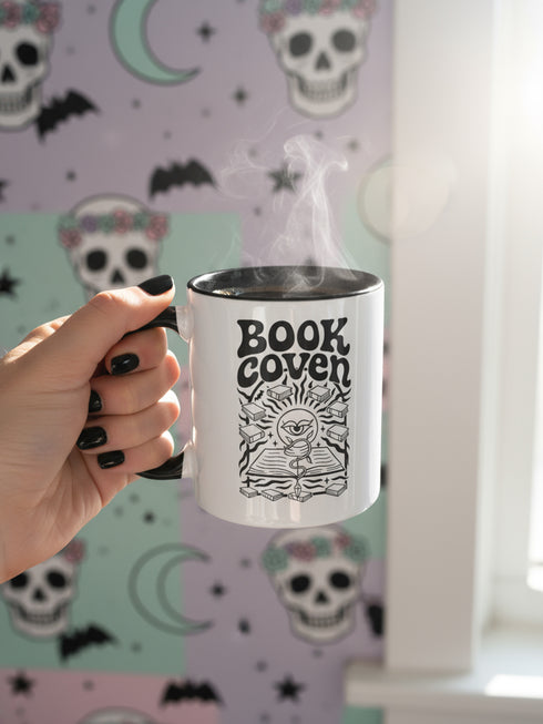 A two-tone ceramic mug with a glossy white exterior and a contrasting black interior, handle, and rim. The front of the mug features a black, rectangular graphic with the words "BOOK COVEN" in a bold, wavy, 70s-inspired retro font. Below the text is an occult-style illustration of an open book on a stand. An all-seeing eye with a snake coiled around it emerges from the book's pages, radiating lines of light. Smaller books float around the central image, completing the witchy, bookish design