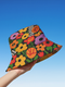 a reversible bucket hat. The visible side features a maximalist 70s-inspired pattern with bold orange, purple, and yellow abstract flowers on a black background with white checkerboard accents. The hat is made of a breathable, linen-feel polyester. The reverse side, hinted at on the brim's underside, has a vibrant Y2K psychedelic print with leopard spots and tropical fruit. This funky, colorful sun hat is a perfect dopamine dressing accessory for festival fashion.