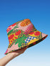 a reversible bucket hat. The visible side features a maximalist 70s-inspired pattern with bold orange, purple, and yellow abstract flowers on a black background with white checkerboard accents. The hat is made of a breathable, linen-feel polyester. The reverse side, hinted at on the brim's underside, has a vibrant Y2K psychedelic print with leopard spots and tropical fruit. This funky, colorful sun hat is a perfect dopamine dressing accessory for festival fashion.