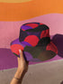 Reversible unisex bucket hat featuring two distinct geometric designs - one side showcases a bold abstract pattern with angular shapes in vibrant purple, pink, red, and black creating dynamic geometric forms and diamond-like structures, while the reverse side displays a contemporary abstract composition with curved organic shapes, linear textures, and color-blocking in purple, red, and black tones. Made from 100% polyester with textured linen feel, this moisture-wicking and breathable hat is available in XS