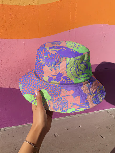 Reversible unisex bucket hat featuring two distinct designs - one side showcases a vibrant floral pop art pattern with purple, pink, green and blue splashes and organic flower motifs, while the reverse side displays a purple giraffe print pattern with dark purple giraffe spots on a light purple background. Made from 100% polyester with textured linen feel, this moisture-wicking and breathable hat is available in XS, S/M, and L/XL sizes for ultimate style versatility.
