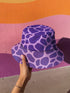 Reversible unisex bucket hat featuring two distinct designs - one side showcases a vibrant floral pop art pattern with purple, pink, green and blue splashes and organic flower motifs, while the reverse side displays a purple giraffe print pattern with dark purple giraffe spots on a light purple background. Made from 100% polyester with textured linen feel, this moisture-wicking and breathable hat is available in XS, S/M, and L/XL sizes for ultimate style versatility.