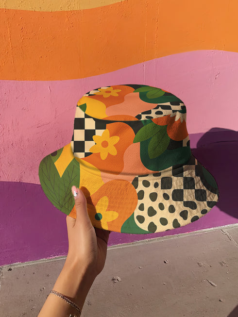 Reversible unisex bucket hat featuring two distinct retro-inspired designs - one side showcases a vibrant 70s-style floral pattern with large orange and yellow flowers, green leaves, and black polka dot accents on cream and coral backgrounds, while the reverse side displays a sophisticated brown and beige floral pattern with orange blooms and neutral tones. Made from 100% polyester with textured linen feel, this moisture-wicking and breathable hat is available in XS, S/M, and L/XL sizes, perfect for vintage