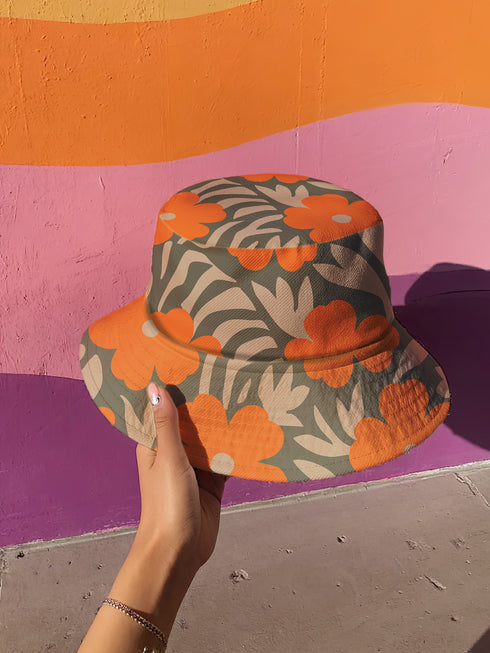 Reversible unisex bucket hat featuring two distinct retro-inspired designs - one side showcases a vibrant 70s-style floral pattern with large orange and yellow flowers, green leaves, and black polka dot accents on cream and coral backgrounds, while the reverse side displays a sophisticated brown and beige floral pattern with orange blooms and neutral tones. Made from 100% polyester with textured linen feel, this moisture-wicking and breathable hat is available in XS, S/M, and L/XL sizes, perfect for vintage