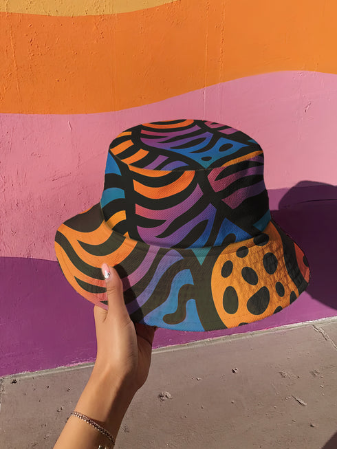 Reversible bucket hat with a bold all-over psychedelic tiger stripe print in pink, blue, yellow, and black; made of moisture-wicking polyester with a linen-texture feel; available in sizes XS, S/M, and L/XL; features two different sides with unique patterns.