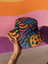 Reversible bucket hat with a bold all-over psychedelic tiger stripe print in pink, blue, yellow, and black; made of moisture-wicking polyester with a linen-texture feel; available in sizes XS, S/M, and L/XL; features two different sides with unique patterns.