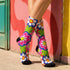 Mid-calf crew sock with a bold 70s psychedelic floral print. The design mixes a red and white diagonal checkerboard with oversized retro flowers, including white daisies with bright yellow centers and a large hot pink bloom with a white inner layer, set over a royal blue background with green leafy foliage. The toe and heel are solid black. The fabric is a soft, stretchy printed knit, made as a one-size unisex statement sock for maximalist and dopamine dressing looks.