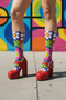Mid-calf crew sock with a bold 70s psychedelic floral print. The design mixes a red and white diagonal checkerboard with oversized retro flowers, including white daisies with bright yellow centers and a large hot pink bloom with a white inner layer, set over a royal blue background with green leafy foliage. The toe and heel are solid black. The fabric is a soft, stretchy printed knit, made as a one-size unisex statement sock for maximalist and dopamine dressing looks.