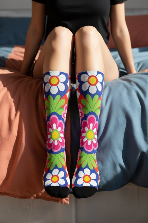 Mid-calf crew sock with a bold 70s psychedelic floral print. The design mixes a red and white diagonal checkerboard with oversized retro flowers, including white daisies with bright yellow centers and a large hot pink bloom with a white inner layer, set over a royal blue background with green leafy foliage. The toe and heel are solid black. The fabric is a soft, stretchy printed knit, made as a one-size unisex statement sock for maximalist and dopamine dressing looks.