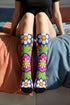 Mid-calf crew sock with a bold 70s psychedelic floral print. The design mixes a red and white diagonal checkerboard with oversized retro flowers, including white daisies with bright yellow centers and a large hot pink bloom with a white inner layer, set over a royal blue background with green leafy foliage. The toe and heel are solid black. The fabric is a soft, stretchy printed knit, made as a one-size unisex statement sock for maximalist and dopamine dressing looks.