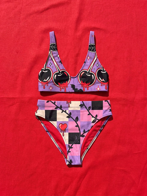 Two piece pastel goth bikini with a v neck top and high waisted bottom. The purple top is covered in large black cherries dripping red, tiny flying bats, and white sparkle stars. The high cut bottoms show a pink, lavender, black, and cream checkerboard overlaid with black thorny vines and dripping red hearts. Soft, stretchy recycled polyester fabric with UPF 50 sun protection and removable padding. Creepy cute whimsigoth swimwear for spooky summer pool days, haunted beach trips, and music festivals.