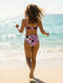 Two piece pastel goth bikini with a v neck top and high waisted bottom. The purple top is covered in large black cherries dripping red, tiny flying bats, and white sparkle stars. The high cut bottoms show a pink, lavender, black, and cream checkerboard overlaid with black thorny vines and dripping red hearts. Soft, stretchy recycled polyester fabric with UPF 50 sun protection and removable padding. Creepy cute whimsigoth swimwear for spooky summer pool days, haunted beach trips, and music festivals.