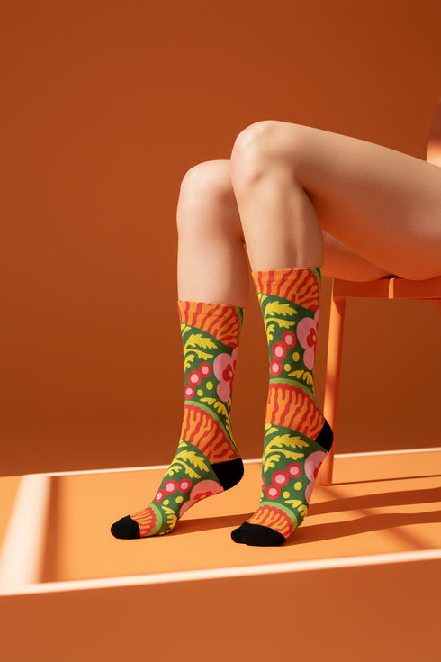 Mid-calf crew sock with a bold retro 70s maximalist botanical print. A deep green base is packed with golden yellow fern-like leaves, pink folk-style flowers with red centers, and clusters of round red and pink berries. Orange and red shell motifs with wavy stripes band the cuff and lower sock. The toe and heel are solid black. The fabric is a soft, stretchy printed knit, made as a one-size unisex statement sock for maximalist and dopamine dressing looks.