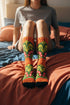 Mid-calf crew sock with a bold retro 70s maximalist botanical print. A deep green base is packed with golden yellow fern-like leaves, pink folk-style flowers with red centers, and clusters of round red and pink berries. Orange and red shell motifs with wavy stripes band the cuff and lower sock. The toe and heel are solid black. The fabric is a soft, stretchy printed knit, made as a one-size unisex statement sock for maximalist and dopamine dressing looks.