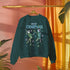 A dark green holiday sweatshirt featuring two dancing skeletons wearing Santa hats. The text "Merry Creepmas" is displayed above them in icy, drippy font. The design is surrounded by colorful snowflakes, blending spooky and festive elements for a fun, alternative holiday look.