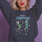 A dark green holiday sweatshirt featuring two dancing skeletons wearing Santa hats. The text "Merry Creepmas" is displayed above them in icy, drippy font. The design is surrounded by colorful snowflakes, blending spooky and festive elements for a fun, alternative holiday look.
