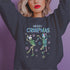 A dark green holiday sweatshirt featuring two dancing skeletons wearing Santa hats. The text "Merry Creepmas" is displayed above them in icy, drippy font. The design is surrounded by colorful snowflakes, blending spooky and festive elements for a fun, alternative holiday look.