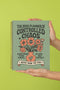A sage green hardcover spiral planner for 2026 with the title "The 2026 Planner of Controlled Chaos" in a funky, retro 70s-style font. The cover features a groovy illustration of orange and pink flowers in a vase, with one flower depicted as an eyeball. The text reads "Bloom where you're planted. Or just wilt artfully." and "Notes from the Void." The planner has a gold metal wire-o binding. The back cover shows a decorative floral frame with a "Property Of" section