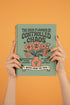 A sage green hardcover spiral planner for 2026 with the title "The 2026 Planner of Controlled Chaos" in a funky, retro 70s-style font. The cover features a groovy illustration of orange and pink flowers in a vase, with one flower depicted as an eyeball. The text reads "Bloom where you're planted. Or just wilt artfully." and "Notes from the Void." The planner has a gold metal wire-o binding. The back cover shows a decorative floral frame with a "Property Of" section