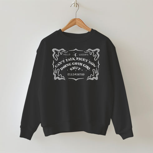 Black sweatshirt featuring white ouija board design with text "CAN'T TALK RIGHT NOW DOING GOTH GIRL SHIT". Ornate border surrounds the design, giving a mystical, witchy vibe.